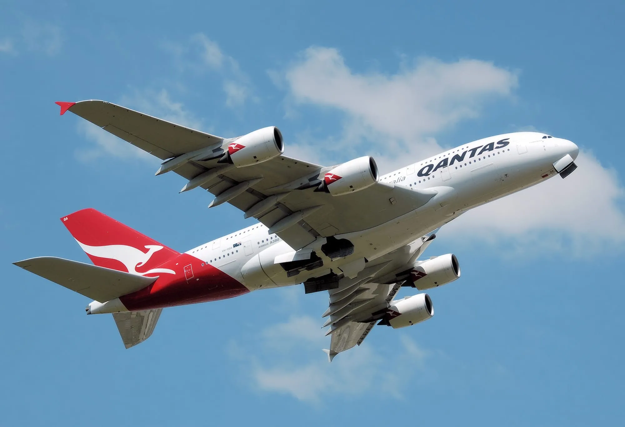 A Qantas Airbus A380 climbing away from the runway against a blue sky, undercarriage doors still open, the flying kangaroo on its red tail.