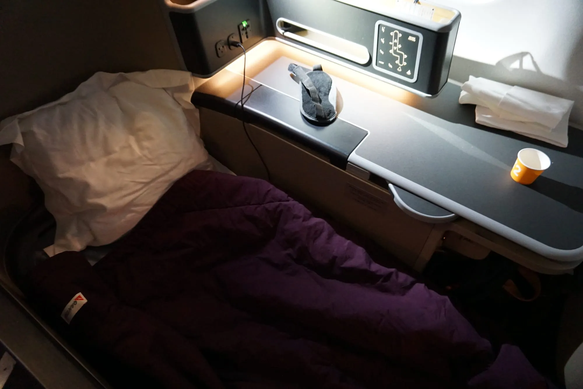 A lie-flat business class seat made up as a bed, pillow and quilt in place, an eye mask on the console and the reading light on.
