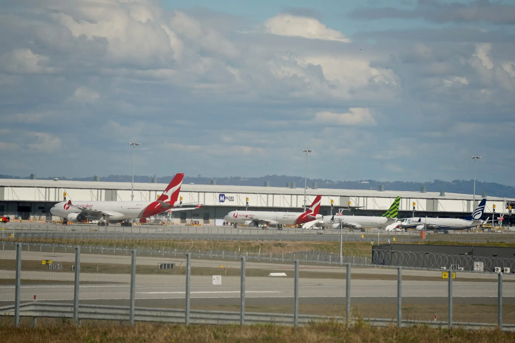 Freighter aircraft in Australia Post, Qantas Freight and Texel Air markings parked in a line outside a long white cargo shed marked Menzies Aviation.